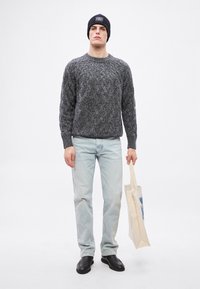 Pelēks adīts džemperis ar rakstu, gaiši zili džinsi, melna cepure ar logotipu, tur rokās bēša auduma somu, un melnas kurpes, stāvot pret vienkāršu fonu.