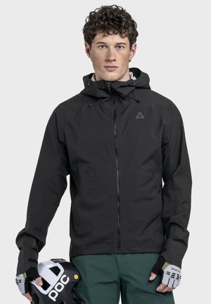 Mann trägt schwarze Kapuzenjacke und grüne Hose, hält schwarzen und weißen POC-Helm mit Radhandschuhen an beiden Händen.