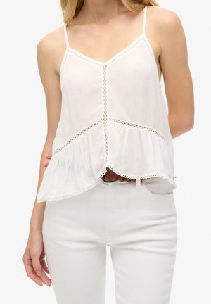 Kobieta ubrana w biały, bezrękawnik z cienkimi ramiączkami, zdobiony koronką i falbaną na dole, w połączeniu z białymi spodniami i brązowym paskiem.