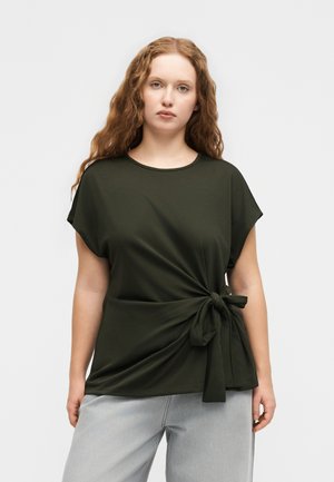 Vrouw met lang, krullend haar die een donkergroene top met korte mouwen draagt, gestrikt in een knoop ter hoogte van de taille, en lichte grijze broek, staand tegen een effen achtergrond.