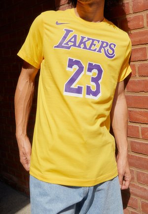 Osoba w żółtej koszulce Lakers z numerem 23 i jasnoniebieskich dżinsach, stojąca przy czerwonej, ceglanej ścianie w świetle słonecznym.