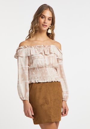 Jonge vrouw met golvend haar draagt een beige blouse met patroon, off-shoulder met ruches en lange mouwen, gecombineerd met een bruine suède minirok.