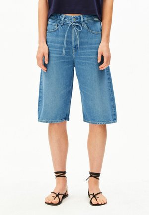 Personne portant un short en denim bleu mi-long avec une taille à cordon, des sandales noires à brides et un haut court foncé.