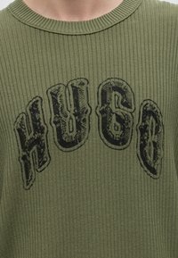 Pull côtelé vert avec de grandes lettres noires usées formant le mot "HUGO" sur la poitrine en arrangement courbe.