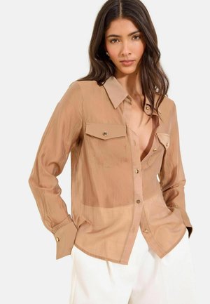 Woman with long dark hair wearing a semi-sheer tan button-up shirt with chest pockets and white pants, hands in pockets, facing forward.