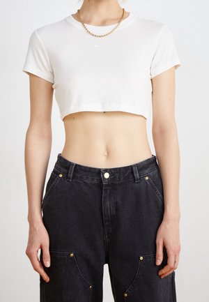 Osoba ubrana w biały krótki T-shirt, złoty łańcuszek oraz wysokie czarne jeansy z kieszeniami i mosiężnymi nitami.