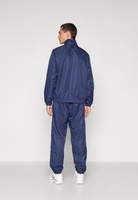 Marineblauer Trainingsanzug aus glattem, synthetischem Material. Die Jacke hat einen hohen Kragen, elastische Bündchen und einen elastischen Bund; die Hose hat elastische Säume.