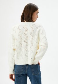 Neulettu kerma sweater, jossa on aaltoiluavoin muotoilu ja ribattu hihansuu, yhdistettynä sinisiin korkeavyötäröisiin farkkuihin. Tekstuuri näyttää pehmeältä ja mukavalta.
