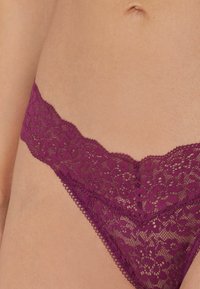 String violet en dentelle avec un motif floral, des bords festonnés et une taille élastique. Texture douce avec un détail de design délicat.