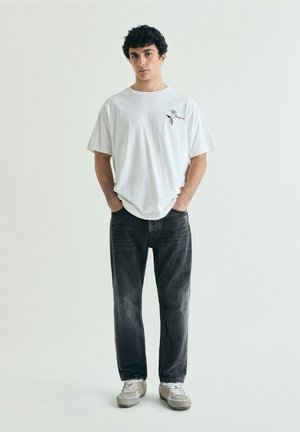 Tânăr în picioare cu mâinile în buzunare, purtând un tricou alb cu grafică, blugi negri estompați și adidași gri pe un fundal simplu.