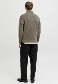 Sweat-shirt vert olive avec col montant, poignets et ourlet côtelés. Porté avec un pantalon noir et des chaussures noires, vu de dos.