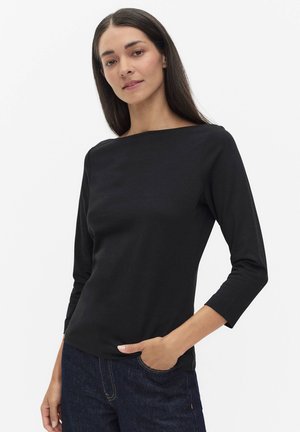 Kvinde med langt mørkt hår iført en sort båd-hals top med trekvart ærmer og mørkeblå jeans, hånd i lommen, neutral baggrund.