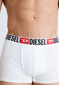 Bijele bokserice s crnim pojasom na kojem se nalazi "DIESEL" logo u crvenoj boji. Tkanina je glatka s prilagođenim oblikom.