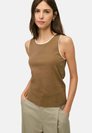 Mujer con pelo largo y castaño que lleva una camiseta marrón sin mangas con ribete blanco y pantalones beige claros, manos en los bolsillos, fondo neutro.