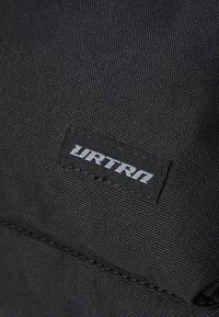 Etiqueta de tela negra con el logo blanco "URTRN", cosida sobre un material negro texturizado, que destaca la durabilidad y los elementos de diseño simples.