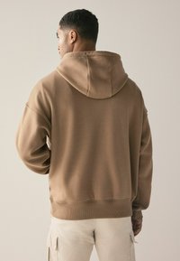 Hnědá mikina s kapucí, volného střihu, s žebrovanými manžetami a spodním lemem. Měkký povrch, minimalistický design bez viditelných log nebo vzorů.