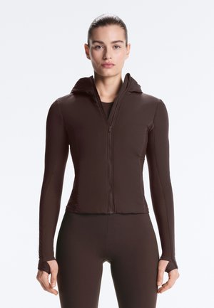 Kobieta ubrana w dopasowaną ciemnobrązową kurtkę z zamkiem i kapturem oraz pasujące legginsy, stojąca na jednolitym jasnym tle.