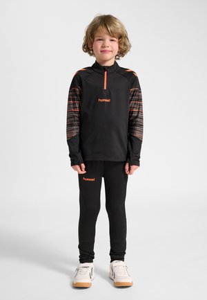 Junges Kind steht, trägt schwarze und orangefarbene Hummel-Sportkleidung mit weißen Turnschuhen, vor einem schlichten hellen Hintergrund.