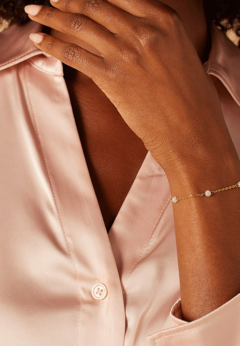 Goldarmband mit kleinen weißen Perlen, getragen an einem Handgelenk, vor einem Hintergrund eines hellrosa Satinhemdes, das am Kragen sichtbar ist.