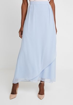 Femme portant une jupe portefeuille bleu clair longueur cheville avec des talons aiguilles couleur nude sur fond blanc.