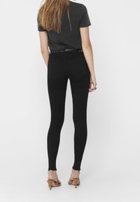 Jeans skinny de tiro alto en negro con una textura suave, que presentan un ajuste ceñido y detalles de costura mínimos, combinados con una camiseta negra y tacones color beige.