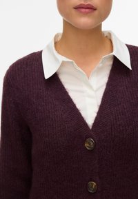Lilla V-hals cardigan laget av teksturert strikkemateriale med tre store brune knapper, brukt over en hvit skjorte med krage.