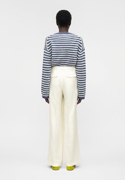 Άτομο που φαίνεται από πίσω, φοράει μπλε και λευκό ριγέ πουλόβερ με μακριά μανίκια, κρεμ παντελόνι με ψηλή μέση και φαρδιά πόδια και κίτρινα παπούτσια.
