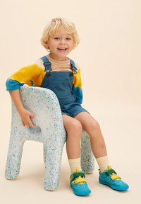 Un bambino è seduto su una sedia bianca maculata, indossando una salopette blu con bretelle regolabili e una maglietta colorata tie-dye. Calzature notevoli in teal e giallo.