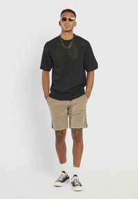 Νεαρός άνδρας φορώντας γυαλιά ηλίου, μαύρο κοντομάνικο πλεκτό πουλόβερ, μπεζ σορτς, λευκές κάλτσες και μαύρα αθλητικά παπούτσια, στέκεται με τα χέρια στις τσέπες.