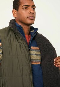 Olivgrüne gesteppte Weste mit hohem Kragen, die über einem gemusterten blau- und cremefarbenen Pullover mit Streifen und einem Fleece-Akzent getragen wird.