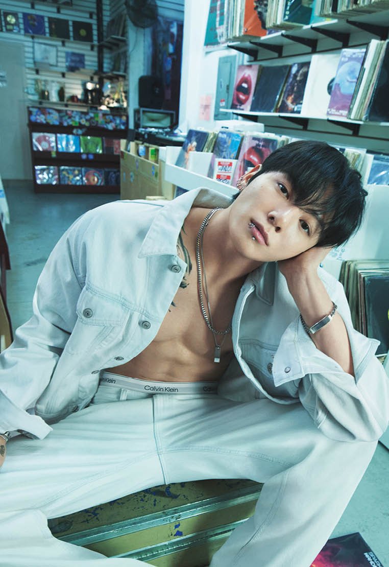 Junger Mann mit Lippenpiercings und mehreren silbernen Halsketten lehnt sich auf seine Hand, trägt eine aufgeknöpfte weiße Jacke und Hose in einem Gang eines Plattenladens.