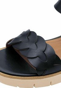 Zwarte leren sandaal met een brede gevlochten band, een tankleurige binnenzool en een stevige beige rubberen zool, met een gladde textuur en minimale details.