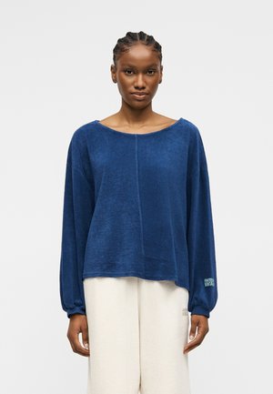 Jeune femme avec des cheveux tressés portant un haut bleu à manches longues ample et un pantalon large beige clair, posant devant un fond uni.