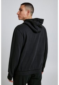 Melnā kapuci džemperis ar brīvu piegriezumu, ribotiem aproču un apakšmalas galiem, un mīkstu tekstūru. Aizmugures skats rāda vienkāršu dizainu bez rotājumiem.