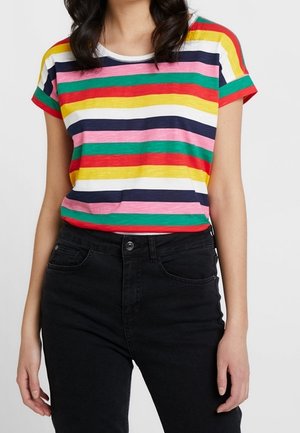 Vrouw draagt een kleurrijk gestreept shirt met korte mouwen, ingestopt in een zwarte high-waisted spijkerbroek tegen een effen achtergrond.