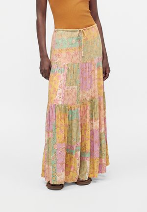 Femme portant une longue jupe patchwork florale multicolore avec un haut orange et des sandales marron, debout sur un fond blanc.
