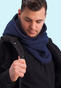 Navy blue knitted scarf featuring a textured rib pattern, worn over a black jacket with a fleece collar. Background is light blue.