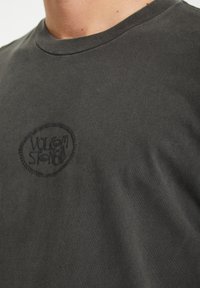 T-shirt grigio scuro realizzato in morbido cotone. Presenta un logo ovale ricamato nero sul petto, con un classico collo a giro e maniche corte.