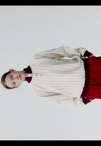 Persona che indossa un pullover bianco a righe sottili con colletto e polsini color granata, abbinato a pantaloni rossi, in piedi contro uno sfondo chiaro uniforme.