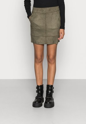 Frau trägt einen olivgrünen Wildleder-Minirock, ein schwarzes Langarmshirt und schwarze klobige Knöchelstiefel mit Schnallen, steht auf einem grauen Boden.