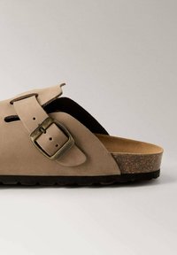 Beige leather sandal featuring a wide strap, adjustable buckle, and a cork footbed, with a textured dark brown sole for grip.