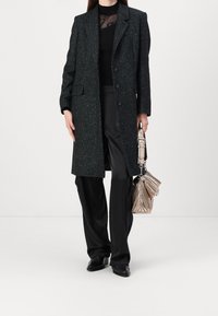 Černý a petrolejově texturovaný kabát s velkými klopami, třemi knoflíky a kapsami, kombinovaný s černými volnými kalhotami a metalickou taškou s záhyby.