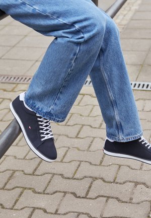 Donkerblauwe sneakers met witte veters en rubberen zolen, met een logo-detail aan de zijkant. Draag ze met lichtblauwe brede jeans.