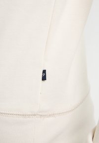 Helles beige Baumwoll-Sweatshirt mit gerippter Struktur und einem kleinen dunkelblauen Etikett mit einem weißen Buchstaben ‚P‘ an der Seitennaht. Nahtdetails sichtbar.