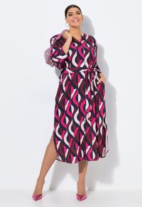 Abito lungo a fantasia nei colori blu navy, rosa e crema, con vita cintata, chiusura a bottoni e spacchi laterali. Abbinato a tacchi alti rosa e una borsa scintillante.
