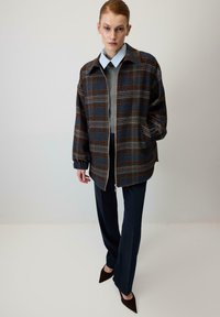 Karirasta jakna v temno modri, rjavi in beli barvi z zadrgo spredaj, prevelikim dizajnom, elastičnimi manšetami, nošena preko sive majice in mornarsko modrih hlač.