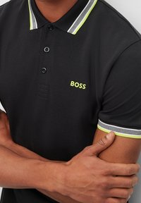 Schwarzes Poloshirt mit kontrastierendem schwarzen und grünen gestreiften Kragen, Logo "BOSS" in Neon-Gelb und strukturiertem Stoff. Kurze Ärmel mit Akzenten.