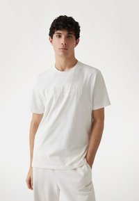Camiseta de algodón blanca con un logo "GUESS" en relieve en la parte frontal. Corte regular con mangas cortas y cuello redondo. Diseño simple.
