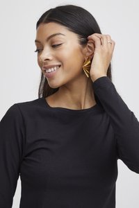 Kvinna med långt mörkt hår som bär en svart topp ler och rör vid sitt guldfärgade fläktformade örhänge med höger hand.