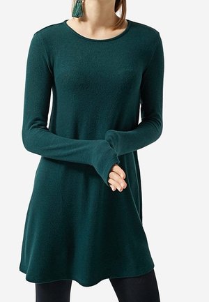 Femme portant une robe en maille vert foncé à manches longues avec un col rond et un legging noir, mains jointes à la taille, sur fond blanc.